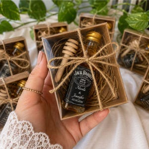 Può includere: Una piccola scatola regalo quadrata in cartone contenente una bottiglia di whisky Jack Daniel's in miniatura e un bastoncino per miele in legno, adagiati su carta marrone sminuzzata. La scatola è legata con spago di juta e ha un coperchio trasparente.