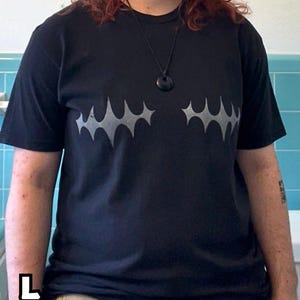 May include: A black short-sleeved t-shirt featuring two symmetrical grey spiky star-like graphic prints across the chest. The shirt is labeled as size L Large. A black cord necklace with a dark circular pendant is worn around the neck.