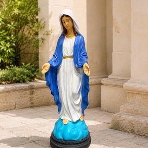 May include: A standing statue of the Virgin Mary wearing a white gown and a blue mantle with a gold-colored belt. The figure has a white head covering, hands extended outward, and stands upon a blue cloud base atop a circular black pedestal.
