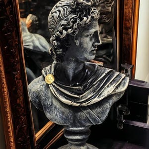 May include: A gray stone bust sculpture of a person with curly hair, wearing a draped garment with a gold accent. The bust is on a pedestal.