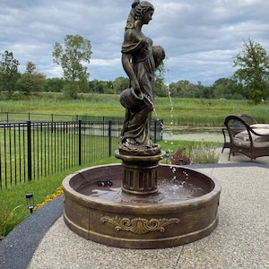 Pode incluir: Uma fonte de cor bronze com uma figura feminina segurando um jarro. A fonte &eacute; cercada por um p&aacute;tio de pedra e uma cerca de metal preto. A figura est&aacute; em p&eacute; sobre um pedestal com uma bacia redonda abaixo. A &aacute;gua flui do jarro para a bacia.