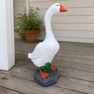 May include: A white goose statue with a bright orange beak and orange webbed feet. The goose is standing on a dark gray, textured rock-like base with a small patch of green grass detail between its feet, captured in a full-body side profile.
