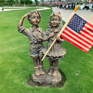 May include: A bronze statue of two children, a boy and a girl, standing on a grassy lawn. The boy is saluting and holding a small object in his hand. The girl is holding a small American flag. The statue is in front of a white building with a crowd of people in the background.