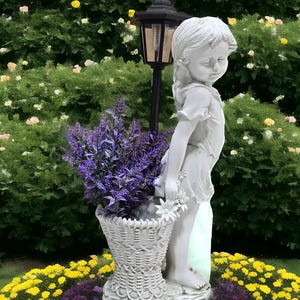 May include: A white stone statue of a girl holding a basket of flowers, set in a garden with yellow and pink flowers. A black lamp post stands behind the statue. The garden is lush with greenery.