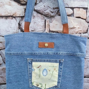 May include: A light blue denim tote bag with brown leather accents and straps. The bag features a decorative pocket with a bird and egg design. The bag is made of denim and has a rectangular shape.