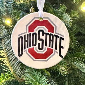 May include: A round, white ceramic ornament with the words "OHIO STATE" in black lettering. The Ohio State University logo is in the center, featuring a red "O" with a gray outline. A white ribbon is attached for hanging.