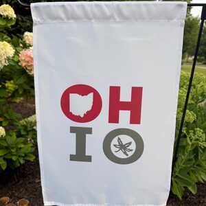 May include: A white rectangular garden flag featuring the text OH IO. The letter O in OH contains a red silhouette of the state of Ohio. The letter H is red. The letter I is gray. The letter O in IO is a gray circle containing a gray buckeye leaf.