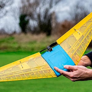 Op de afbeelding: Een op afstand bestuurd vliegend vleugelvliegtuig met een felgele, doorschijnende vleugelstructuur, een centrale blauwe romp en een kleine zwarte propeller gemonteerd op het achterste bovendeel van het hoofdgedeelte.