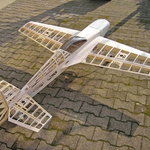 Puede incluir: Un modelo de avión de madera blanca con una cabina transparente. El modelo está en estado de montaje parcial, con las alas y la cola unidas, pero el fuselaje aún está abierto. El modelo está sentado sobre una superficie de hormigón gris.