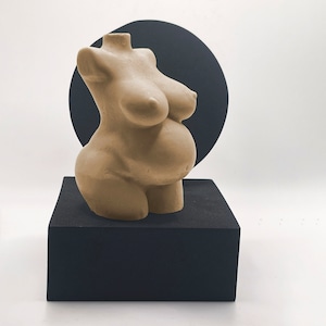 May include: A beige-colored, pregnant torso sculpture. The sculpture is displayed on a black square pedestal, with a black circular backdrop. The sculpture has a smooth texture and detailed anatomical features.