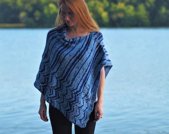 May include: A woman wearing a blue and white striped knitted poncho with a chevron pattern. She is standing in front of a lake with a wooded background.