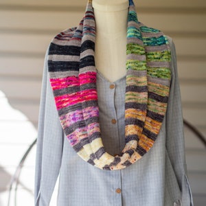 May include: A colorful striped infinity scarf made of yarn in shades of grey, black, pink, purple, yellow, orange, and green.