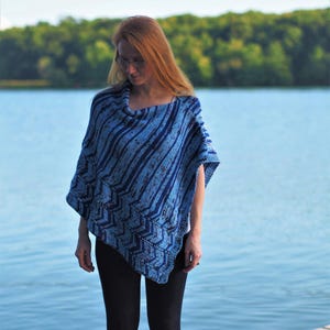 May include: A woman wearing a blue and white striped knitted poncho with a chevron pattern. She is standing in front of a lake with a wooded background.