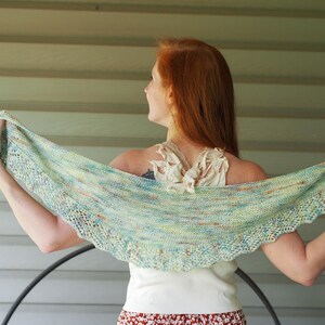 May include: A light blue and green shawl with a scalloped edge. The shawl is held up, showcasing its intricate knit pattern and color variations. The shawl is a soft, lightweight accessory.