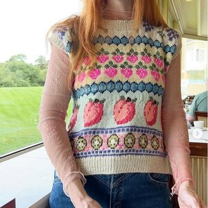 May include: A cream-colored knit vest with a strawberry pattern in pink, blue, and green. The vest is worn over a sheer pink long-sleeved top and paired with blue jeans. The vest features a round neckline and a sleeveless design.