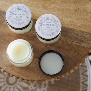 May include: Overhead shot of three small jars of castor oil salve on a wooden surface. Two jars are closed, displaying a white label with floral accents and the text "Castor Oil Salve." One jar is open, revealing the salve's pale yellow color.