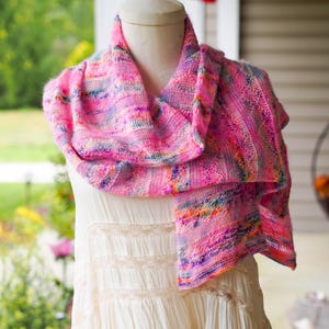 May include: A vibrant pink and multi-colored knitted scarf draped over a mannequin. The scarf features a blend of pink, purple, blue, and yellow hues. The scarf is displayed over a cream-colored dress. The Cape May Fiber Co. logo is in the top left corner.