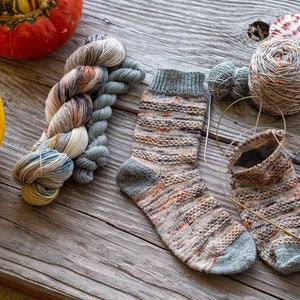 May include: A pair of knitted socks in a multi-colored yarn with a grey heel and toe. The socks are on a wooden surface with yarn balls, knitting needles, and seashells.