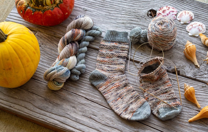 May include: A pair of knitted socks in a multi-colored yarn with a grey heel and toe. The socks are on a wooden surface with yarn balls, knitting needles, and seashells.