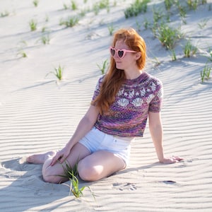 May include: A woman wearing a colorful knit sweater with a seashell design sits on a sandy beach. She is wearing white shorts and pink heart-shaped sunglasses.