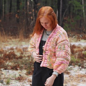 May include: A chunky knit cardigan in shades of pink, yellow, and green. The open-front sweater has a textured design with ribbed sleeves. The model is wearing a gray shirt and black overalls. The background is a forest scene.