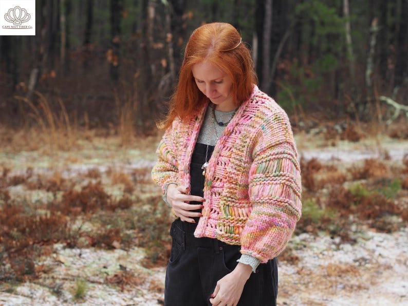 May include: A chunky knit cardigan in shades of pink, yellow, and green. The open-front sweater has a textured design with ribbed sleeves. The model is wearing a gray shirt and black overalls. The background is a forest scene.