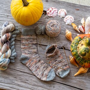 May include: Two pairs of hand-knitted socks in a colorful yarn with a speckled pattern. The socks are laid out on a wooden surface with a pumpkin, yarn balls, and seashells.