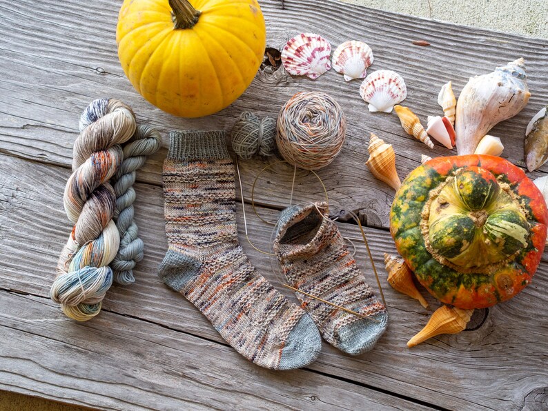 May include: Two pairs of hand-knitted socks in a colorful yarn with a speckled pattern. The socks are laid out on a wooden surface with a pumpkin, yarn balls, and seashells.