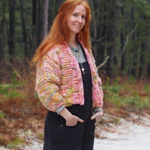 May include: A woman wearing a chunky, hand-knitted pink and yellow cardigan. The cardigan has a textured, open weave design. She is wearing black overalls and a light green shirt. The background is a forest with tall trees.