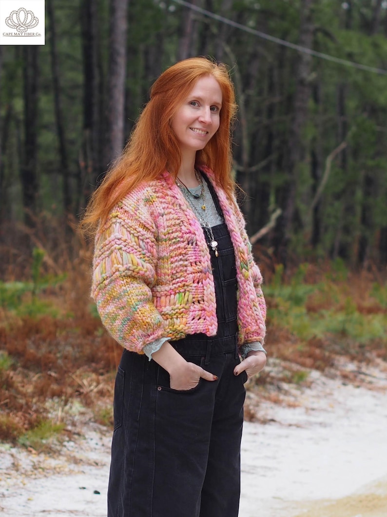 May include: A woman wearing a chunky, hand-knitted pink and yellow cardigan. The cardigan has a textured, open weave design. She is wearing black overalls and a light green shirt. The background is a forest with tall trees.