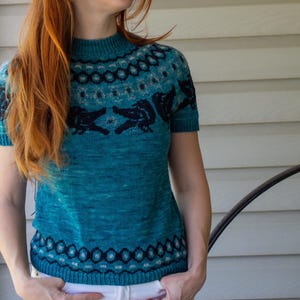 May include: A short-sleeved, teal-colored knit sweater featuring a mock neck and a dark blue, intricate geometric and animal-inspired pattern around the yoke and the hemline. The garment is constructed with a ribbed collar, cuffs, and bottom hem.