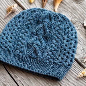 Könnte beinhalten: Eine blaue Strickmütze mit Zopfmuster. Die Mütze ist auf einer Holzoberfläche mit Muscheln fotografiert.