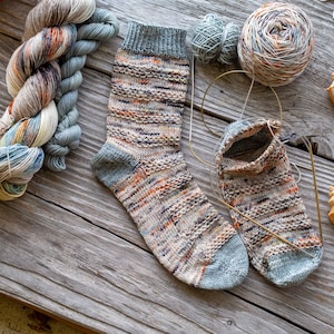 May include: A pair of hand-knitted socks in a blue, brown, and beige color scheme. The socks are lying on a wooden surface with yarn and seashells.
