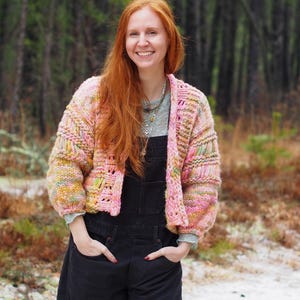 May include: A multi-colored, chunky knit cardigan in shades of pink, yellow, and green. The open-front sweater has a boxy shape with long sleeves. The person in the image is wearing black overalls.