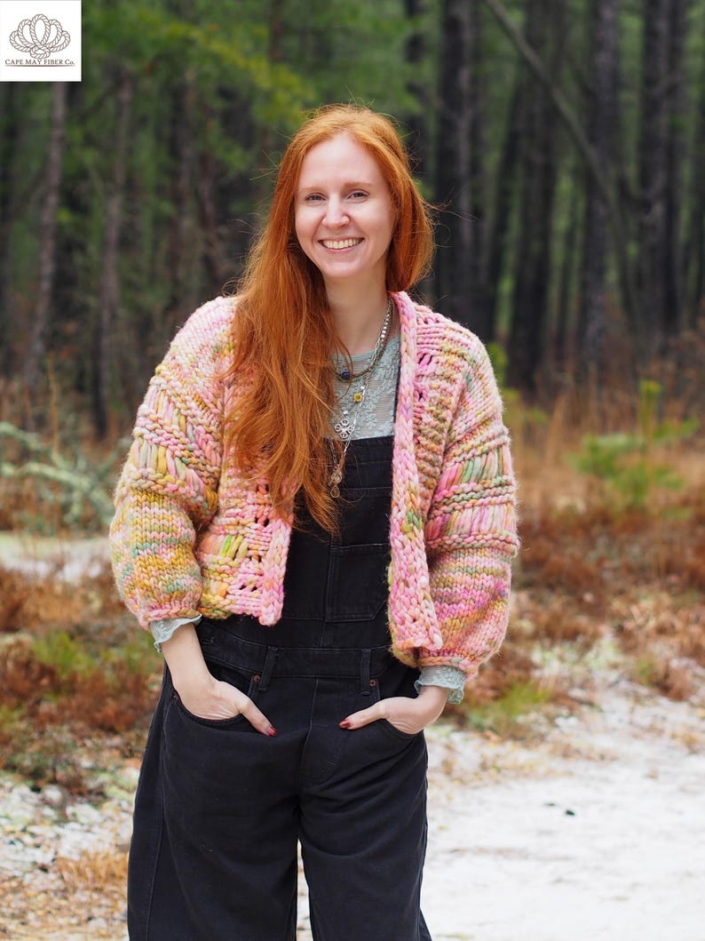 May include: A multi-colored, chunky knit cardigan in shades of pink, yellow, and green. The open-front sweater has a boxy shape with long sleeves. The person in the image is wearing black overalls.