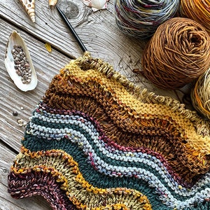 Könnte beinhalten: Nahaufnahme einer gestrickten Schals mit einem wellenförmigen Muster in Braun-, Grün-, Blau- und Gelbtönen. Der Schal wird mit Stricknadeln bearbeitet und Wollknäuel in verschiedenen Farben sind im Hintergrund sichtbar.