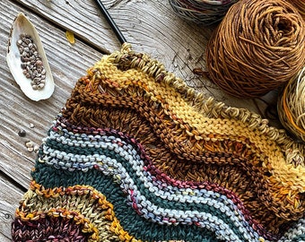 May include: A close-up of a knitted scarf with a wavy pattern in shades of brown, green, blue, and yellow. The scarf is being worked on with knitting needles and yarn balls in various colors are visible in the background.