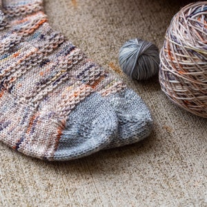 May include: A pair of knitted socks in a multi-colored yarn with a grey heel and toe. The socks are lying on a textured surface next to two balls of yarn.
