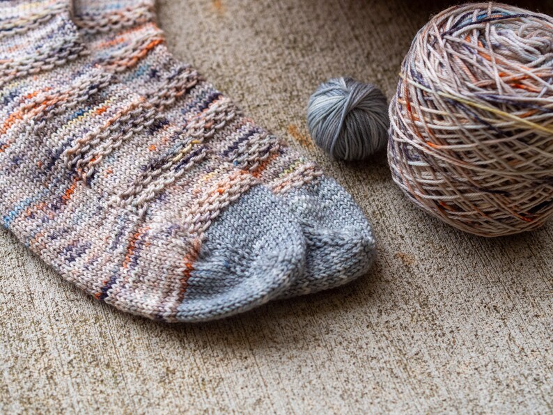 May include: A pair of knitted socks in a multi-colored yarn with a grey heel and toe. The socks are lying on a textured surface next to two balls of yarn.