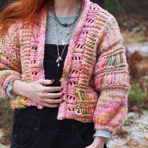 May include: A chunky, open-front cardigan in a vibrant mix of pink, yellow, and green. The hand-knitted sweater features a textured stitch pattern and voluminous sleeves. The garment is approximately 24 inches long.