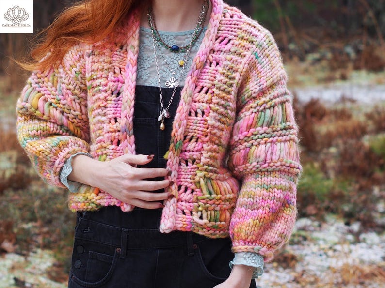 May include: A chunky, open-front cardigan in a vibrant mix of pink, yellow, and green. The hand-knitted sweater features a textured stitch pattern and voluminous sleeves. The garment is approximately 24 inches long.