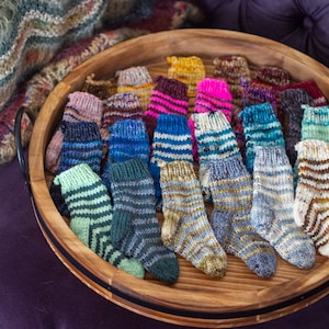 Op de afbeelding: Een houten dienblad gevuld met kleurrijke handgebreide sokken. De sokken zijn in verschillende patronen en kleuren, waaronder blauw, groen, roze en bruin.