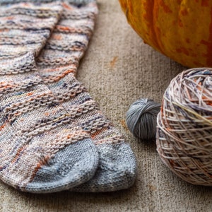 May include: A pair of hand-knitted socks in a colorful, variegated yarn with a grey base. The socks are lying on a textured, beige surface with a pumpkin and yarn balls nearby.