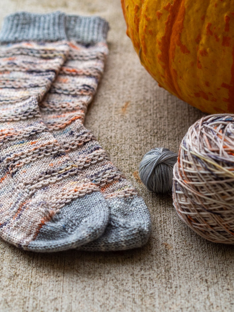 May include: A pair of hand-knitted socks in a colorful, variegated yarn with a grey base. The socks are lying on a textured, beige surface with a pumpkin and yarn balls nearby.