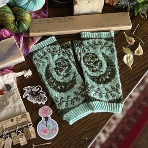 May include: A pair of teal green knitted fingerless gloves with a brown and green pattern. The gloves are laid out on a wooden surface with other items, including a box of incense sticks, a small bag, and a few decorative items.