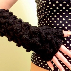 May include: A black knitted fingerless glove with a textured design. The glove is worn on the left hand of a person wearing a black and white polka dot top.