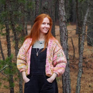 May include: A woman wearing a pink, yellow, and green knit cardigan over black overalls. The cardigan has a textured, open-weave design. The woman is smiling in a forest setting.
