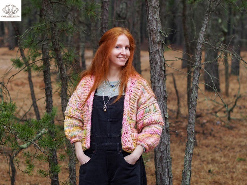 May include: A woman wearing a pink, yellow, and green knit cardigan over black overalls. The cardigan has a textured, open-weave design. The woman is smiling in a forest setting.