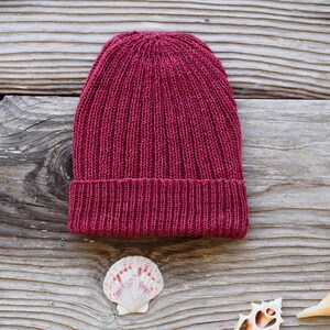 May include: A knitted burgundy beanie hat with a ribbed brim. The hat is displayed on a weathered wooden surface, with seashells scattered around it. The hat is a solid color and appears to be handmade.