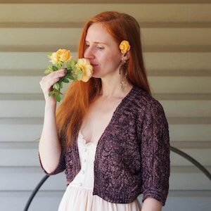 Puede incluir: Una mujer con un bolero de punto morado oscuro con un estampado floral. Sostiene rosas amarillas y tiene una rosa amarilla en el pelo. El suéter está sobre un vestido blanco. El fondo es una pared de color claro.